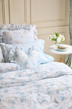 a bed with blue and white floral comforter on it's side table next to a window