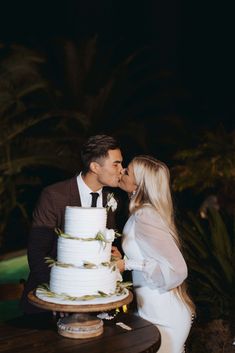 a man and woman kissing in front of a white wedding cake with greenery on top