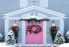 a pink door with wreaths and lights in front of a house covered in snow