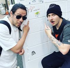 two men standing next to each other near a white brick wall with graffiti on it