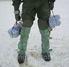 a man in green pants and boots holding two water bottles on the snow covered ground