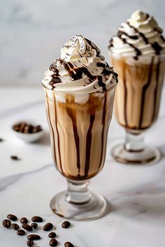 two glasses filled with hot chocolate and whipped cream on top of coffee beans next to each other