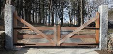 an open wooden gate with stone pillars leading to a driveway in front of some trees