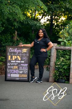 a woman standing next to a chalkboard sign in front of some bushes and trees