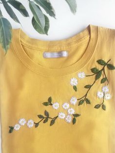 a yellow t - shirt with white flowers and green leaves on the front, next to a potted plant