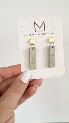 a pair of earrings is being held up in front of a card with the letter m on it