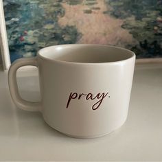 a white coffee cup with the word pray written on it sitting in front of a painting