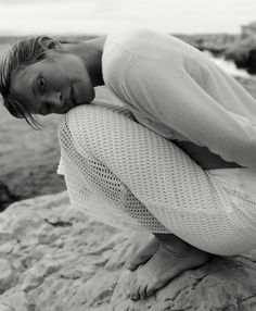 a black and white photo of a woman sitting on rocks by the ocean with her arms around her head