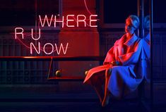 a woman sitting on a bench in front of a neon sign that says, where ru now