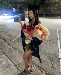 a woman dressed as a graduate holding flowers and a bouquet in her hand while standing on the sidewalk