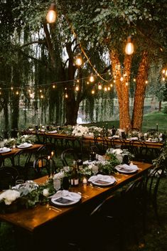 an outdoor dinner table set up with lights strung from the trees and greenery on the tables