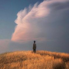 a man standing on top of a dry grass covered hill under a cloudy blue sky