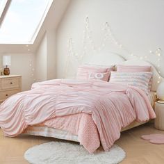 a bed with pink comforter and pillows in a white room next to a window