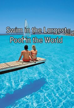 two people sitting on a dock in the middle of blue water with a sailboat