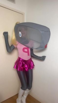 a woman in a pink shirt and silver skirt standing next to a wall with a large object on it's head