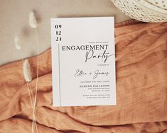 an engagement party card sitting on top of a bed next to some dried flowers and a straw hat