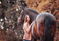 a woman standing next to a brown horse in front of a tree filled forest with lots of leaves