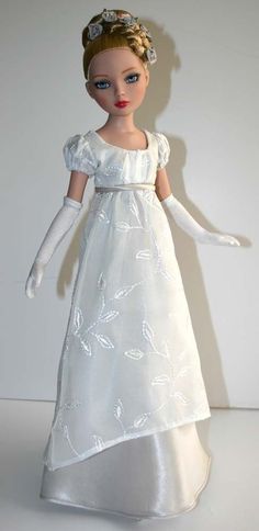 a doll in a white dress and gloves on a white surface with a wall behind it