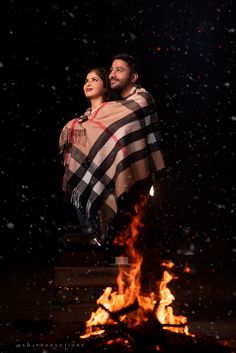 a man and woman wrapped in a blanket sitting on top of a fire with snow falling around them