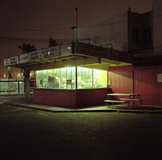 an empty street corner at night with a lit up store front and bench in the foreground