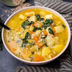 a white bowl filled with soup and vegetables on top of a gray table next to a spoon