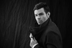 black and white photograph of a man in a coat looking at the camera with his hand on his tie