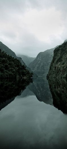 a body of water surrounded by mountains under a cloudy sky with trees in the foreground