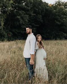 a pregnant woman standing next to a man in a field with tall grass and trees