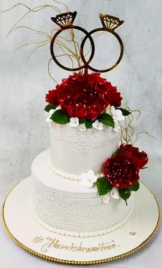 a wedding cake decorated with red flowers and two gold rings on top, sitting on a white plate