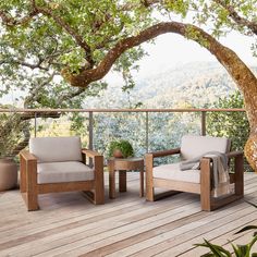 two chairs and a table on a wooden deck under a tree with mountains in the background