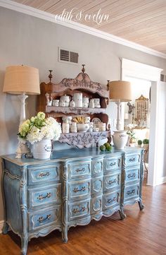 a blue dresser with flowers and vases on it in front of a wall mounted china cabinet