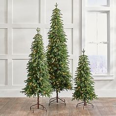 three artificial christmas trees with lights on them in front of a white wall and wooden floor