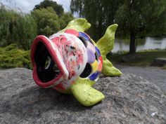 a colorful ceramic fish sculpture sitting on top of a rock next to a body of water