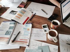 a wooden table topped with lots of papers next to a laptop computer and coffee cup