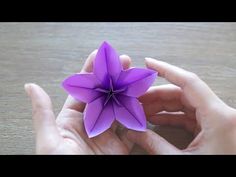 two hands holding a purple origami flower on top of a wooden table next to another person's hand