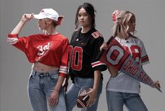 three girls in sports jerseys and baseball caps are posing for the camera, one girl is holding a football bat