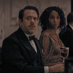 a man in a tuxedo sitting next to a woman with a glass of wine