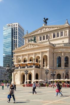 people walking around in front of a large building with statues on it's sides