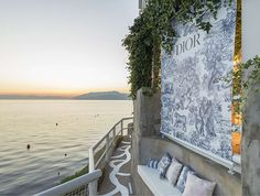 an outdoor seating area overlooks the ocean at sunset, with greenery growing on the wall