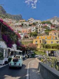 an old car is driving down the road in front of some buildings and flowers on the hillside