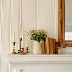 a white mantle with books, candles and flowers on it in front of a mirror