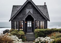 a black house sitting on top of a lush green hillside next to the ocean with white flowers