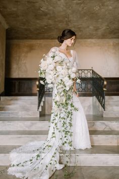 a woman in a wedding dress holding a bouquet of white flowers and greenery standing on some steps