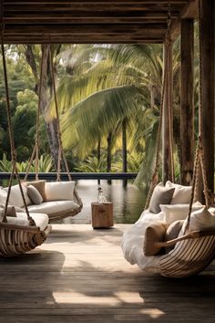 two hanging chairs on a wooden deck next to a body of water with palm trees in the background