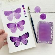 a person holding a purple butterfly next to some watercolor paints and a stamp pad