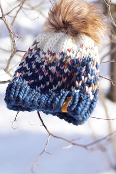 a knitted hat hanging from a tree branch in the snow with a brown fur pom - pom