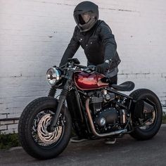 a man in black jacket and helmet sitting on a red motorcycle parked next to a white brick wall