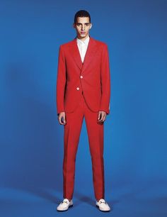 a young man in a red suit and white shirt standing against a blue background with his hands in his pockets