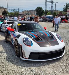 two sports cars parked on gravel with people standing around them in front of some buildings