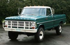 an old green truck parked in a parking lot next to some trees and bushes on a rainy day